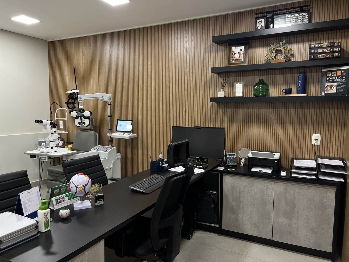 Consultório OFTA VITTA, mesa de atendimento, painel ripado de madeira e prateleiras com literatura médica